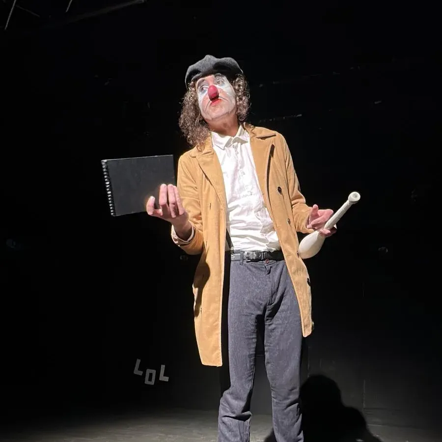 JYE in clown on a dark stage, holding a notebook and a juggling club.